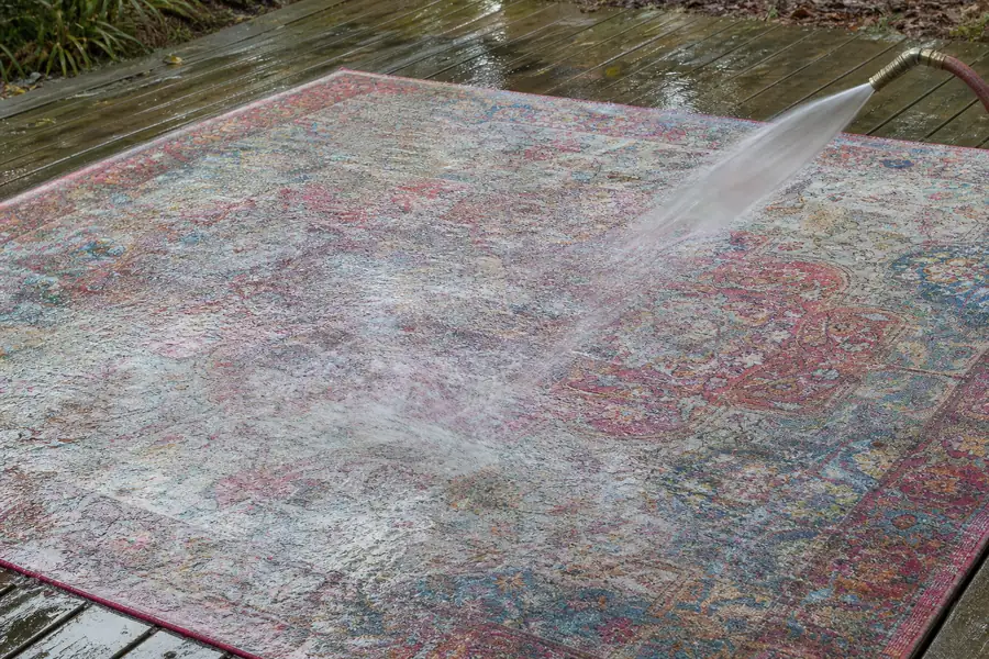 Colorful area rug laid flat on a wooden deck being rinsed with a garden hose