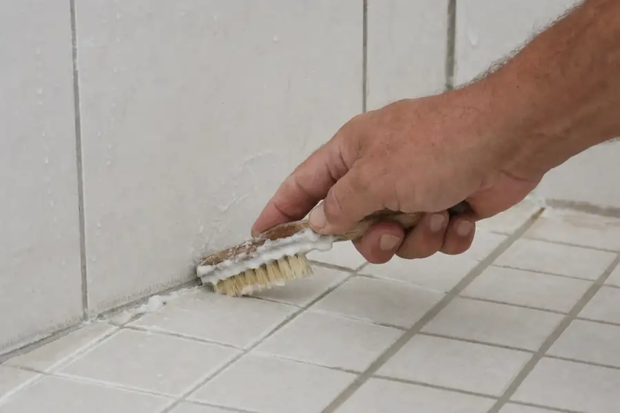 Hand scrubbing shower grout lines with a stiff brush and thick baking soda paste