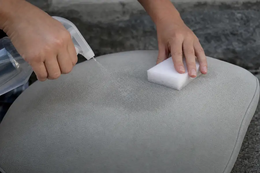 Spraying rubbing alcohol onto a microfiber cushion while scrubbing with a white sponge