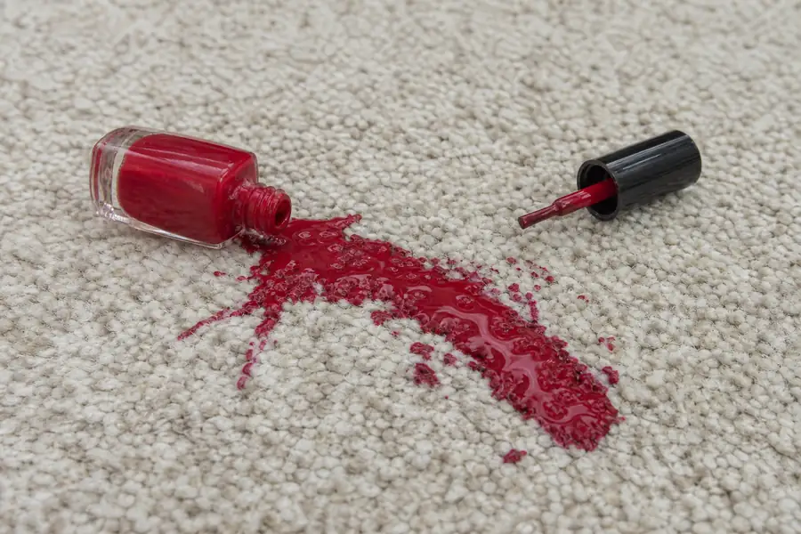 Open red nail polish bottle tipped over on light carpet with a fresh spill