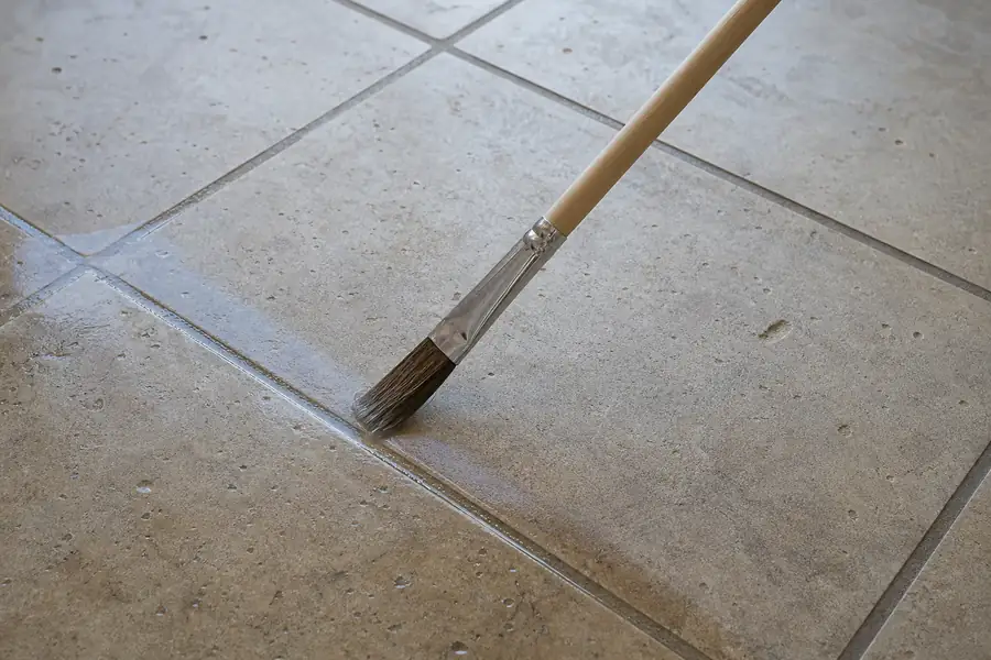 Applicator brush drawing clear sealer along grout lines of a tiled floor